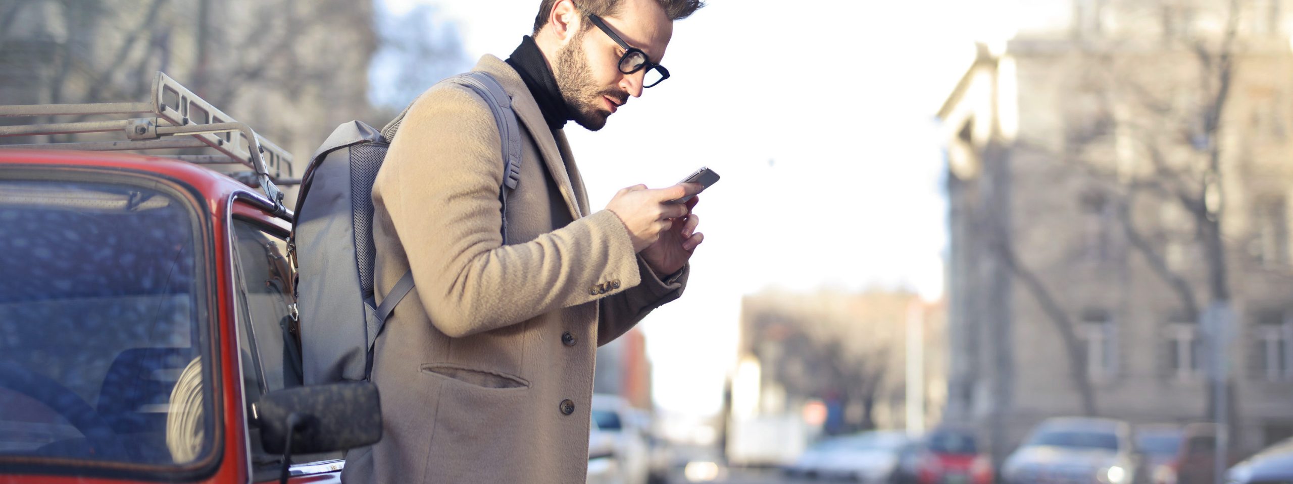 Man in Beige Coat Holding Phone Leaning on Red Vehicle